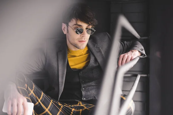 Enfoque selectivo del hombre guapo en gafas de sol sentado en las escaleras y sosteniendo el teléfono inteligente en la mano - foto de stock