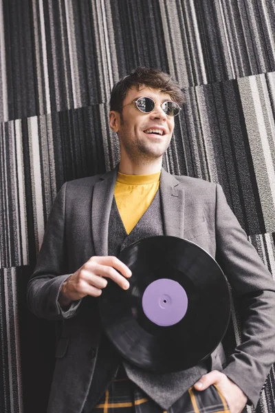 Cheerful young man in sunglasses holding plastic retro vinyl record and standing with hand in pocket — Stock Photo