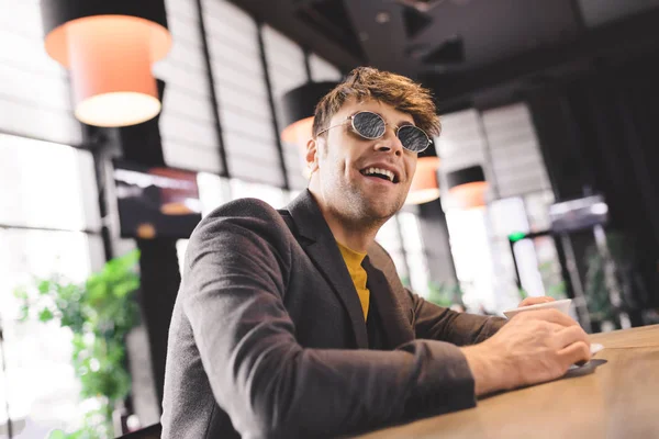 Hombre guapo en gafas de sol sonriendo cerca de la taza con café mientras está sentado en el mostrador del bar - foto de stock
