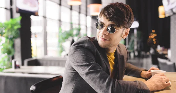 Joven alegre en gafas de sol sonriendo mientras está sentado en el mostrador del bar cerca de la taza con café - foto de stock