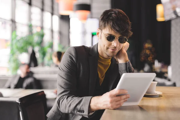 Fröhlicher junger Mann mit Sonnenbrille sitzt am Tresen und hält ein digitales Tablet in der Nähe der Tasse mit Kaffee — Stockfoto