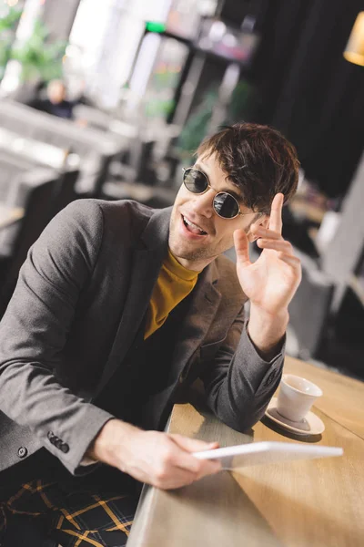 Щасливий молодий чоловік в сонцезахисних окулярах жестикулює біля барної стійки, тримаючи цифровий планшет біля чашки з кавою — стокове фото