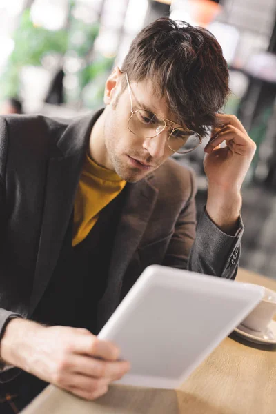 Thoughtful young man in glasses sitting at bar counter while looking at digital tablet near cup with coffee — Stock Photo
