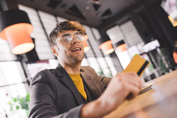 Bel homme dans des lunettes assis au comptoir du bar tout en tenant la carte de crédit dans le café — Photo de stock