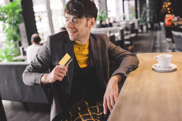 Hombre alegre en vasos sentado en el mostrador del bar mientras sostiene la tarjeta de crédito en la cafetería - foto de stock