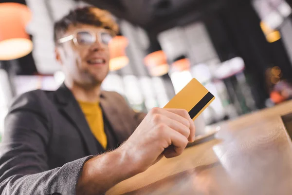 Selective focus of credit card in hand of cheerful man in glasses at bar counter — Stock Photo