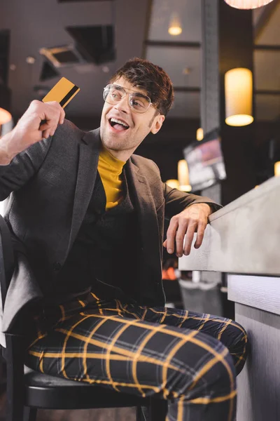 Hombre excitado en vasos sentado en el mostrador del bar mientras sostiene la tarjeta de crédito en la cafetería - foto de stock