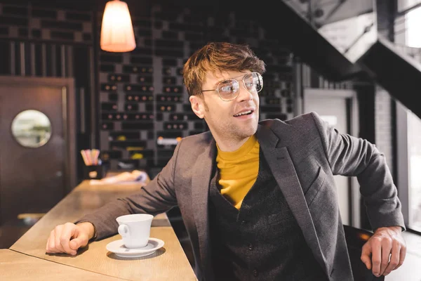 Красивый молодой человек в очках сидит за барной стойкой возле чашки кофе — стоковое фото