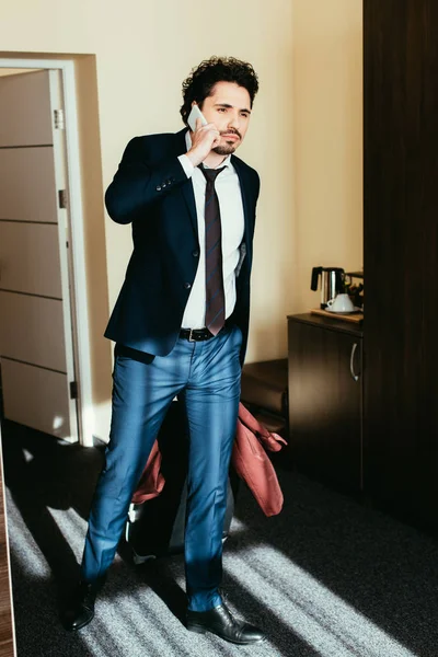 Бизнесмен в костюме разговаривает на смартфоне в гостиничном номере с чемоданом — стоковое фото