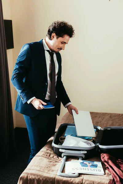 Homme d'affaires titulaire d'un passeport et mettre un ordinateur portable dans la valise sur le lit dans la chambre d'hôtel — Photo de stock