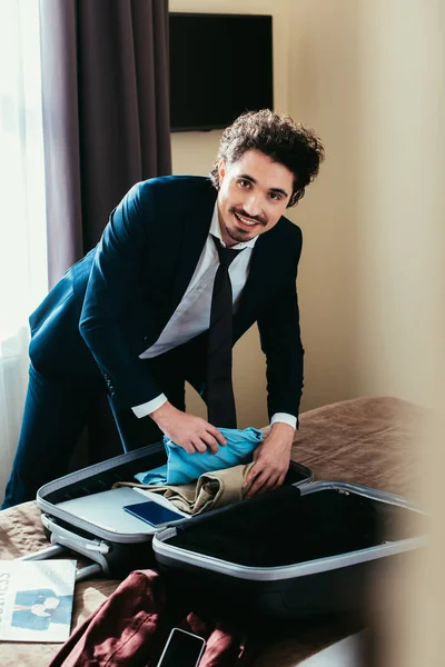 Hombre de negocios sonriente con ropa en bolsa de viaje en la cama en la habitación de hotel - foto de stock