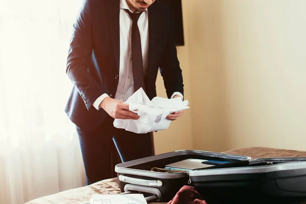 Cropped view of adult businessman putting clothes in suitcase on bed in hotel room — Stock Photo
