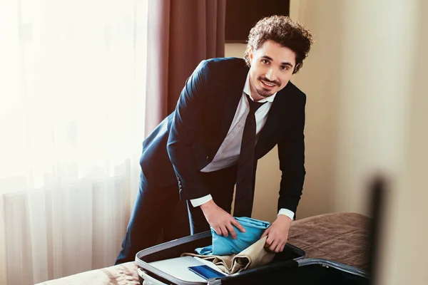 Красивый бизнесмен с одеждой и паспортом в чемодане на кровати в номере отеля — стоковое фото