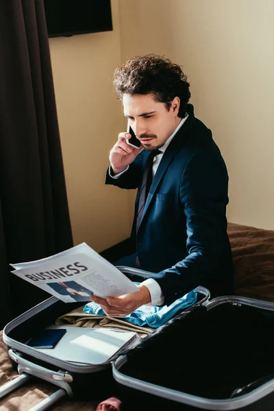 Hombre de negocios hablando en el teléfono inteligente y mirando el periódico de negocios mientras está sentado en la cama con la maleta en la habitación de hotel - foto de stock
