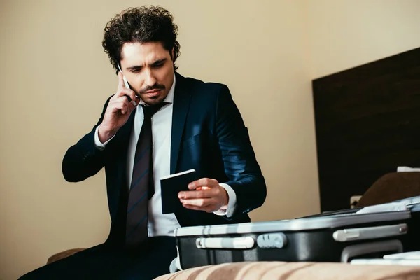 Hombre de negocios guapo hablando en el teléfono inteligente y la celebración de pasaporte mientras está sentado en la cama con el equipaje en la habitación de hotel - foto de stock