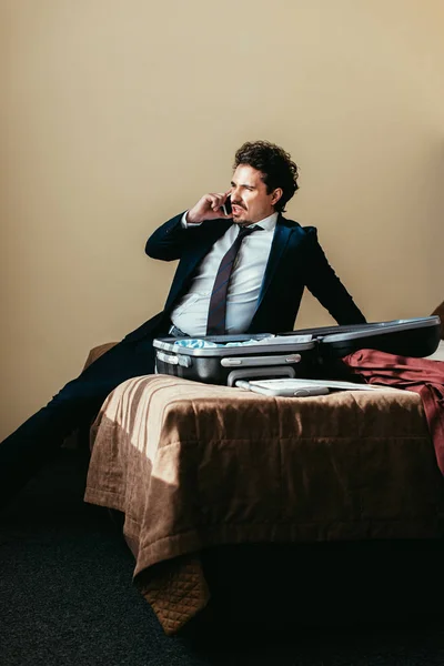 Businessman in suit talking on smartphone while sitting on bed with suitcase in hotel room — Stock Photo