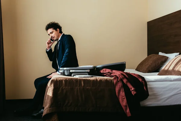 Homem de negócios falando no smartphone enquanto sentado na cama com mala no quarto de hotel — Fotografia de Stock