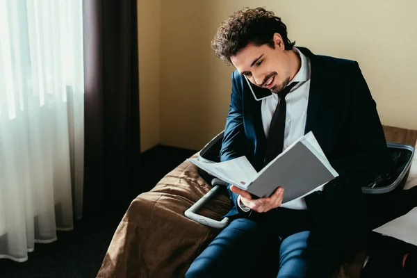 Hombre de negocios hablando en el teléfono inteligente y mirando los documentos en la habitación de hotel - foto de stock