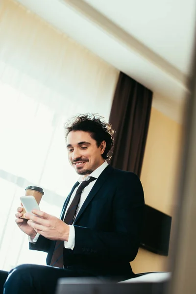 Homem de negócios sorrindo usando smartphone e segurando café para ir no quarto de hotel — Fotografia de Stock