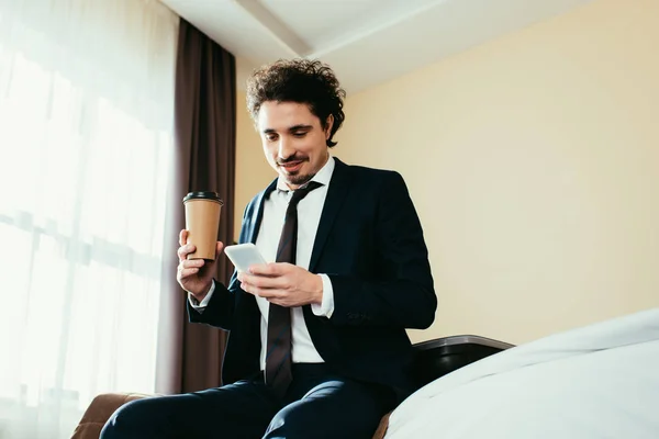 Happy businessman using smartphone and holding disposable cup of coffee in hotel room — Stock Photo
