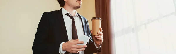 Vue recadrée de l'homme d'affaires en utilisant un smartphone et en tenant du café pour aller dans la chambre d'hôtel avec la lumière du soleil — Photo de stock