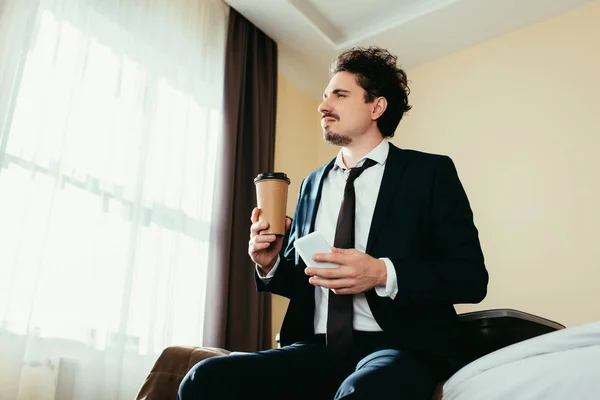 Hombre de negocios pensativo utilizando el teléfono inteligente y la celebración de café para ir en la habitación de hotel - foto de stock