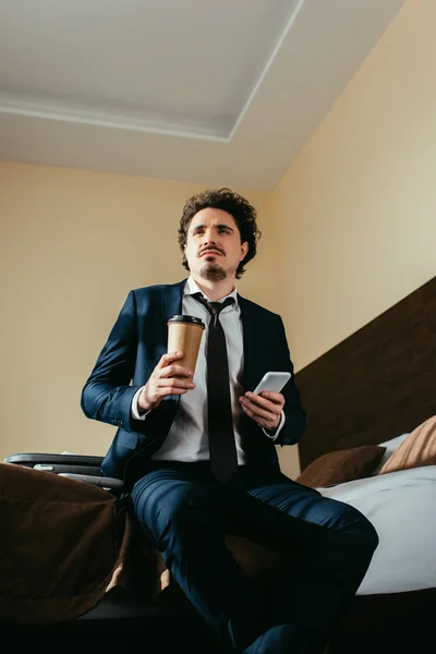 Pensativo hombre de negocios utilizando el teléfono inteligente y la celebración de café para ir en la habitación de hotel - foto de stock