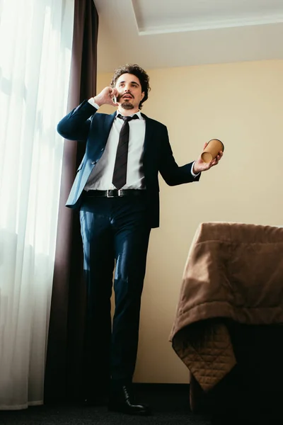 Бизнесмен в костюме разговаривает на смартфоне и держит кофе, чтобы пойти в гостиничный номер — стоковое фото