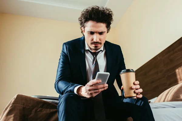 Molesto hombre de negocios utilizando el teléfono inteligente y la celebración de la taza de café desechable en la habitación de hotel - foto de stock