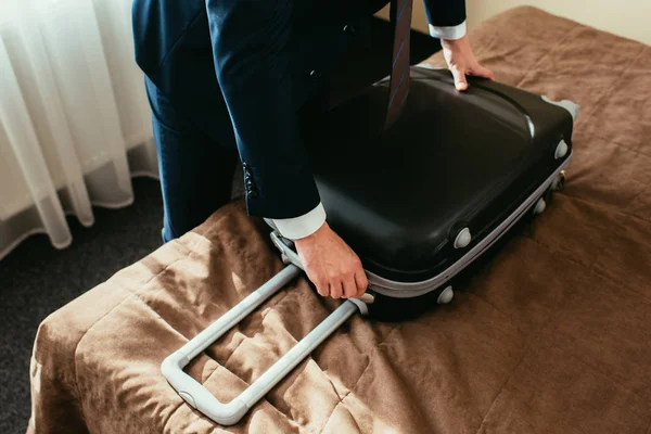 Vista cortada de homem de negócios em terno com mala na cama no quarto de hotel — Fotografia de Stock