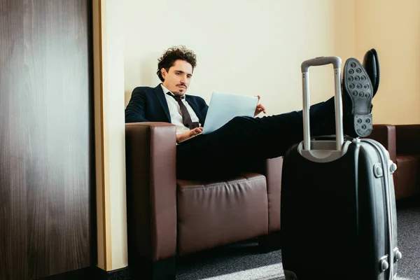 Uomo d'affari telelavoro su laptop con gambe sulla borsa da viaggio in camera d'albergo — Foto stock