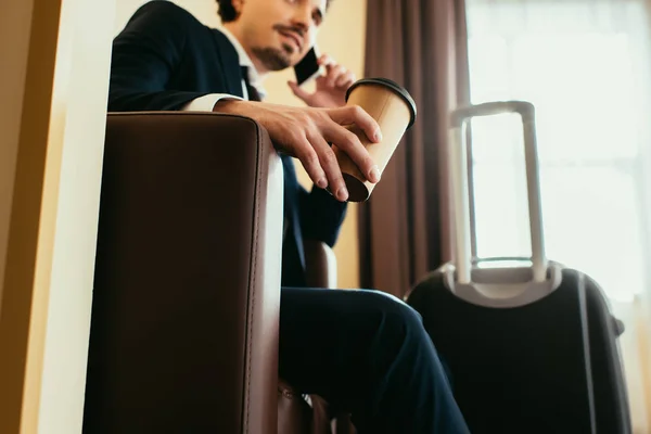 Businessman with disposable cup of coffee talking on smartphone in hotel room with suitcase — Stock Photo