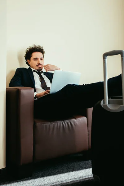 Бізнесмен у костюмі, що працює на ноутбуці з ногами на валізі в готельному номері — стокове фото