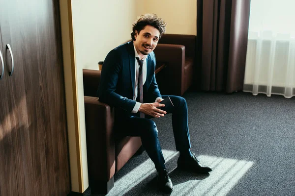 Homem de negócios feliz segurando passaporte e sentado no quarto de hotel — Fotografia de Stock