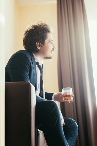 Hombre de negocios soñador con traje sosteniendo un vaso de whisky en la habitación de hotel - foto de stock
