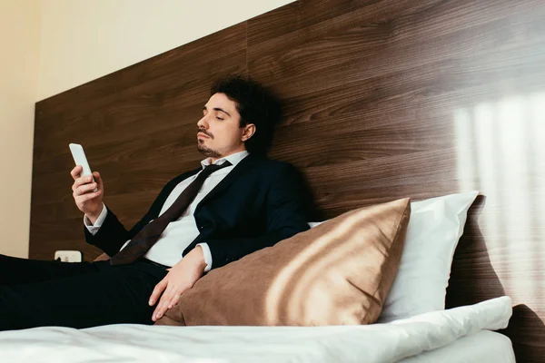 Hombre de negocios en traje usando teléfono inteligente en la cama en la habitación de hotel - foto de stock