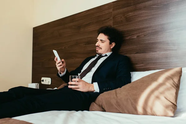 Hombre de negocios adulto usando teléfono inteligente y sosteniendo vaso de bebida alcohólica en la cama en la habitación de hotel - foto de stock