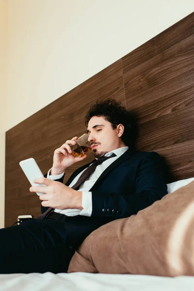 Businessman using smartphone and drinking whiskey on bed in hotel room — Stock Photo