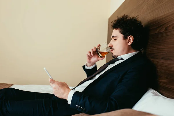 Schöner Geschäftsmann benutzt Smartphone und trinkt Whiskey auf Bett im Hotelzimmer — Stockfoto