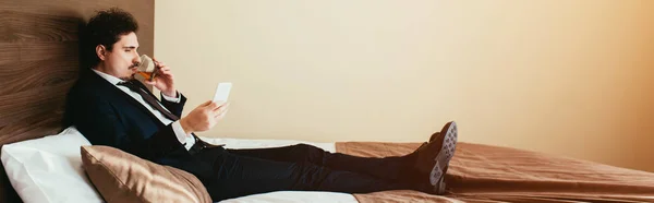 Бизнесмен в костюме с помощью смартфона и питьевой виски на кровати в номере отеля — стоковое фото
