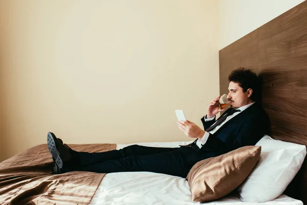 Hombre de negocios en ropa formal con teléfono inteligente y beber whisky en la cama en la habitación de hotel - foto de stock