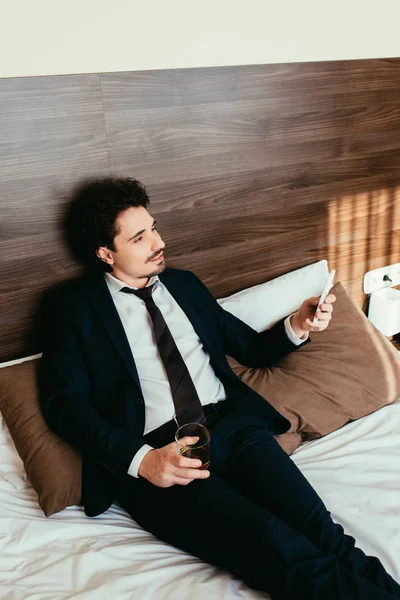 Hombre de negocios usando teléfono inteligente y sosteniendo el vaso de coñac en la cama en la habitación de hotel - foto de stock