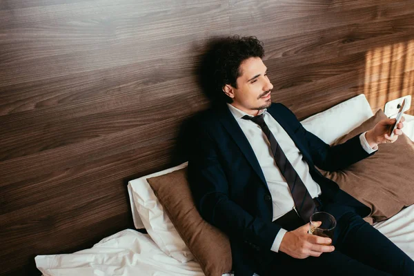 Hombre de negocios usando un smartphone y sosteniendo un vaso de whisky en la cama en la habitación de hotel - foto de stock