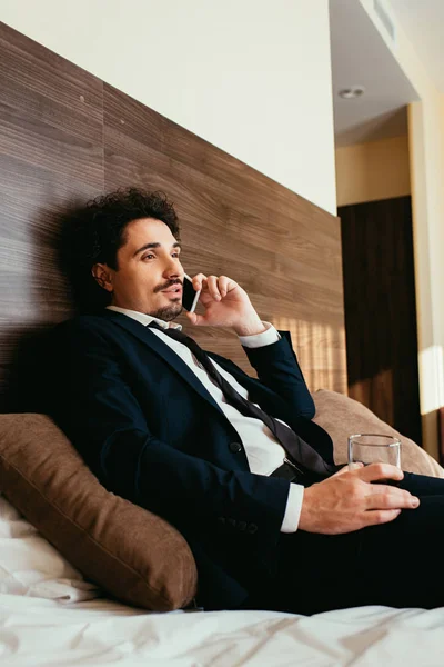 Hombre de negocios hablando en el teléfono inteligente y sosteniendo vaso de whisky en la cama en la habitación de hotel - foto de stock