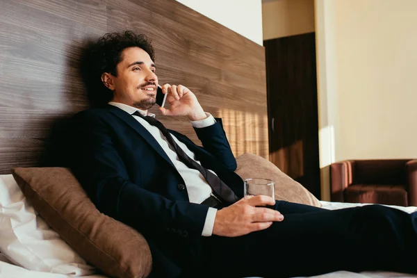 Hombre de negocios sonriente hablando en un teléfono inteligente y sosteniendo un vaso de whisky en la cama en la habitación del hotel - foto de stock