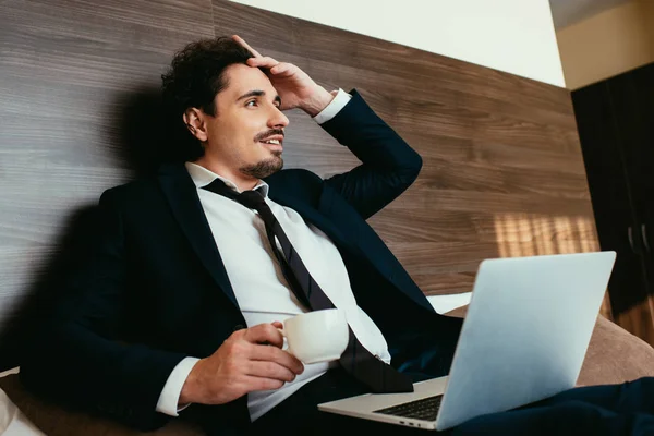 Bel homme d'affaires en costume tenant une tasse de café et utilisant un ordinateur portable dans la chambre d'hôtel — Photo de stock