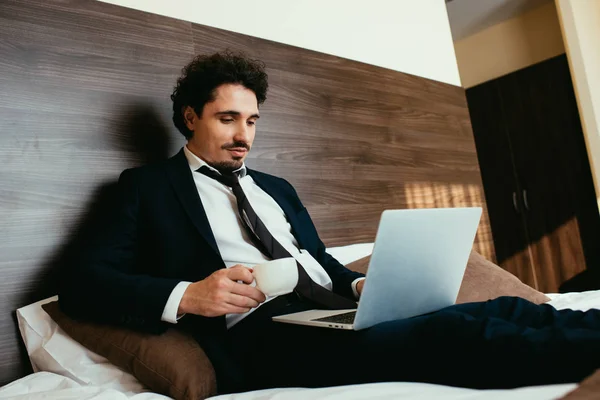Geschäftsmann im Anzug hält Tasse Kaffee in der Hand und arbeitet im Hotelzimmer am Laptop — Stockfoto