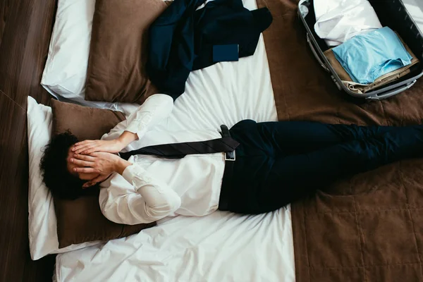 Vista superior del hombre de negocios agotado descansando en la cama con bolsa de viaje en la habitación de hotel - foto de stock