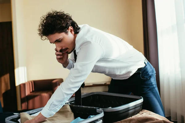 Hombre de negocios guapo hablando en el teléfono inteligente mientras se pone la ropa en la bolsa de viaje - foto de stock