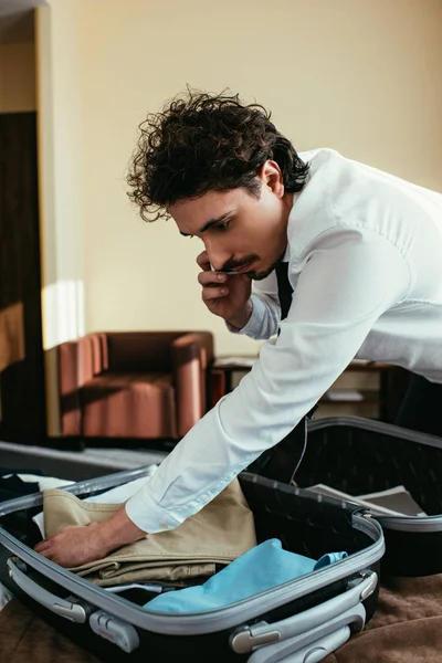 Businessman putting clothes into travel bag while talking on smartphone — Stock Photo